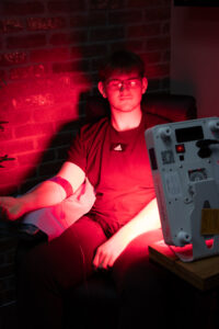 A man sits comfortably in a chair with an IV drip inserted into his arm, receiving hydration therapy. A red light panel glows behind him, casting a warm red hue. He appears relaxed, with a calm expression, as the IV infusion and red light therapy work simultaneously.